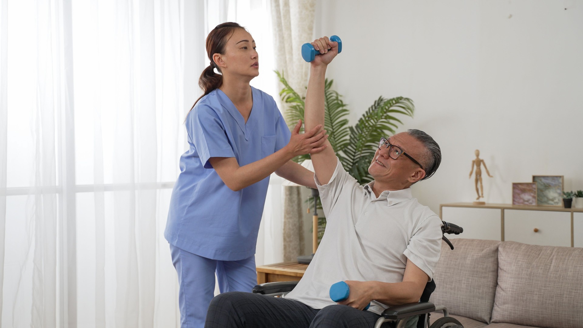 asian female nurse assisting disabled grandfather on wheelchair while he is holding up dumbbells for rehab training at home. domiciliary care for senior people concept